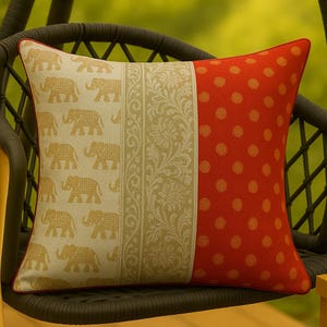 May include: Square decorative pillow with a red border. The pillow features three distinct sections: a cream-colored section with gold elephant patterns, a beige floral patterned section, and a red section with gold polka dots. The pillow is on a dark brown chair.