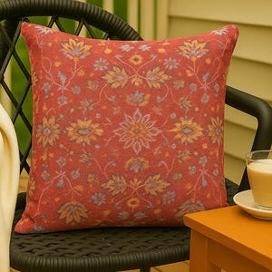 May include: A square decorative pillow with a floral pattern in shades of red, yellow, and blue. The pillow is placed on a black wicker chair. A small orange table and a glass of a beverage are also visible.