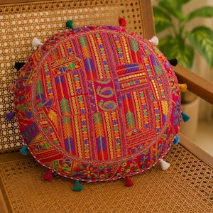 May include: Round decorative cushion with a vibrant red background and intricate embroidered patterns in various colors. The cushion features a detailed design with paisley and geometric shapes, and is trimmed with colorful tassels.