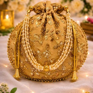 Peut inclure: Un sac potli doré orné de broderies dorées complexes, de paillettes et d'une fermeture à cordon. Le sac est agrémenté de multiples rangs de fausses perles blanches servant de poignée, complétés par des pompons métalliques dorés à la base.