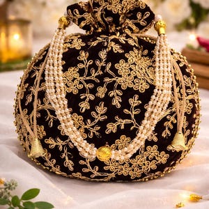 May include: A round, black velvet clutch with gold floral embroidery. The bag features a drawstring closure and is adorned with a pearl and gold beaded strap. Gold tassels and a gold coin add to the elegant design.
