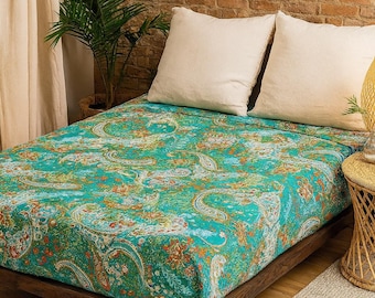 May include: A bed with a teal paisley patterned bedspread and two white pillows. The bed frame is made of dark wood. A small plant and a woven side table are visible.