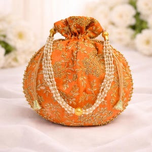 May include: A round, orange pouch with gold floral embroidery and a drawstring closure. It features a beaded pearl necklace and gold tassels. The background is a soft white fabric.