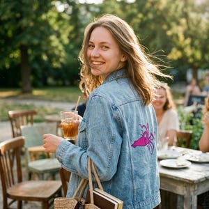 Könnte beinhalten: Eine hellblaue Jeansjacke mit einer leuchtend violetten Tintenfischgrafik auf der Rückseite. Die Jacke verfügt über einen klassischen Kragen, lange Ärmel mit geknöpften Manschetten und sichtbare Nähte entlang der Einsätze und Taschen.