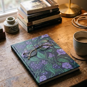 Könnte beinhalten: Ein Notizbuch mit einem floralen Muster aus lila Blumen und grünen Blättern, mit einer Brille darauf. Ein Stapel Bücher, eine Tasse Kaffee und ein Tintenfass befinden sich ebenfalls auf der Holzoberfläche.