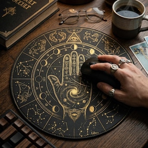 Puede incluir: Una alfombrilla de ratón circular negra con un diseño celestial en color dorado. El gráfico incluye una palma central con una galaxia en espiral, rodeada de fases lunares, constelaciones del zodiaco y un símbolo de ojo. La palabra Libra aparece abajo.