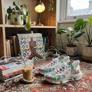 May include: A pair of colorful sneakers with a vibrant pattern of pink, blue, and yellow shapes. The shoes have white laces and a clear sole. The image also includes a vinyl record, a glass of iced coffee, and potted plants.
