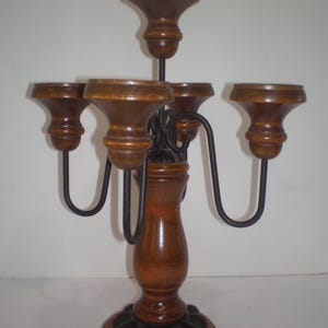 May include: A five-arm candelabra featuring a central wooden pillar and base with a dark brown finish. Four curved black metal arms extend from the center, each topped with a wooden candle cup. A fifth wooden candle cup sits atop the central pillar.
