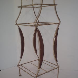 May include: A tall, tiered metal stand featuring a gold-toned wire frame with two square shelves. The structure is accented by three curved, dark brown wooden side panels that connect the upper and lower sections, supported by four thin metal legs.