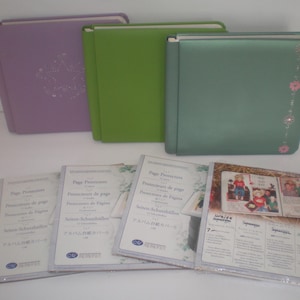 May include: Three square photo albums in purple, green, and teal, with a set of clear page protectors. The purple album has a star design. The teal album has pink flower accents. The page protectors have text in multiple languages.