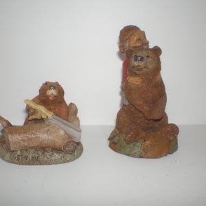 May include: Two brown figurines. One depicts a beaver sawing a log, with the word "OVERTIME" on the base. The other shows a person hugging a bear. Both are detailed with realistic textures and colors.