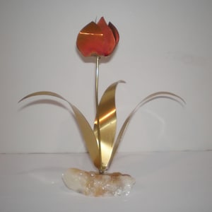 May include: A decorative metal flower sculpture featuring a copper-colored tulip bloom atop a thin brass stem. The flower has three long, curved brass leaves extending from the base, which is anchored in a rough, translucent white quartz stone.