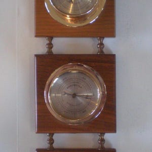 May include: A wooden wall-mounted weather station with three circular gauges. Each gauge has a brass-colored bezel and a silver-colored face. The top unit has a ring for hanging. The wood is a medium brown color.