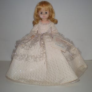 May include: A vintage doll with blonde hair and blue eyes, wearing a cream-colored dress with silver trim and a tag that reads "Cinderella". The doll's dress has a sparkly, textured pattern.