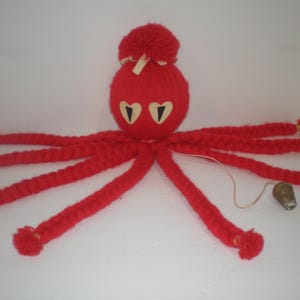May include: A red yarn octopus decoration featuring a round head with a pom-pom top, cream-colored ribbon accents, and two heart-shaped eyes with black pupils. Eight long, braided yarn tentacles extend outward, each finished with a small red pom-pom.