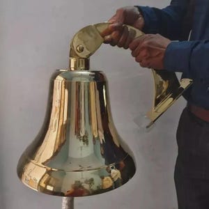 Puede incluir: Una gran campana de latón brillante con un asa curva. La campana tiene una base acampanada y una cuerda adjunta. La campana está siendo sostenida por una persona.