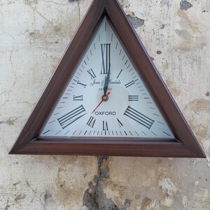 Puede incluir: Un reloj de pared triangular con un marco de madera marrón oscuro. La esfera del reloj es blanca con números romanos y manecillas negras. El texto "Jones & Hendricks EST 1923 OXFORD" está impreso en la esfera. El segundero es rojo.