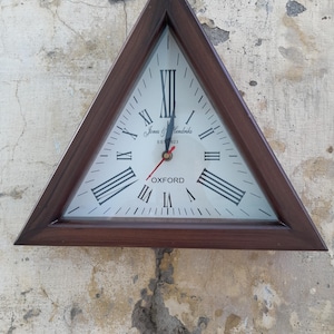 Puede incluir: Un reloj de pared triangular con un marco de madera marrón oscuro y una esfera blanca. El reloj presenta números romanos negros, manecillas de hora y minuto negras y un segundero rojo. El texto Jones & Hendriks EST 1923 y OXFORD está impreso en la esfera.