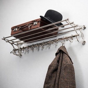 Puede incluir: Un perchero de pared cromado pulido con un estante, ganchos y adornos decorativos. Una maleta de cuero marrón y un sombrero bombín negro descansan sobre el estante. Un abrigo de tweed marrón cuelga de uno de los ganchos.