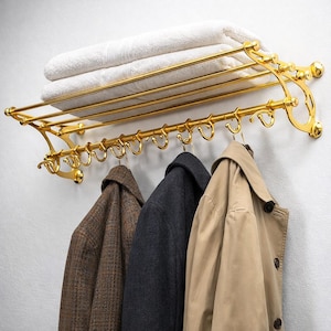 Puede incluir: Un estante dorado montado en la pared con un estante que sostiene toallas blancas dobladas y una fila de ganchos debajo. Tres abrigos en marrón, gris y beige cuelgan de los ganchos. El estante tiene un diseño ornamentado.