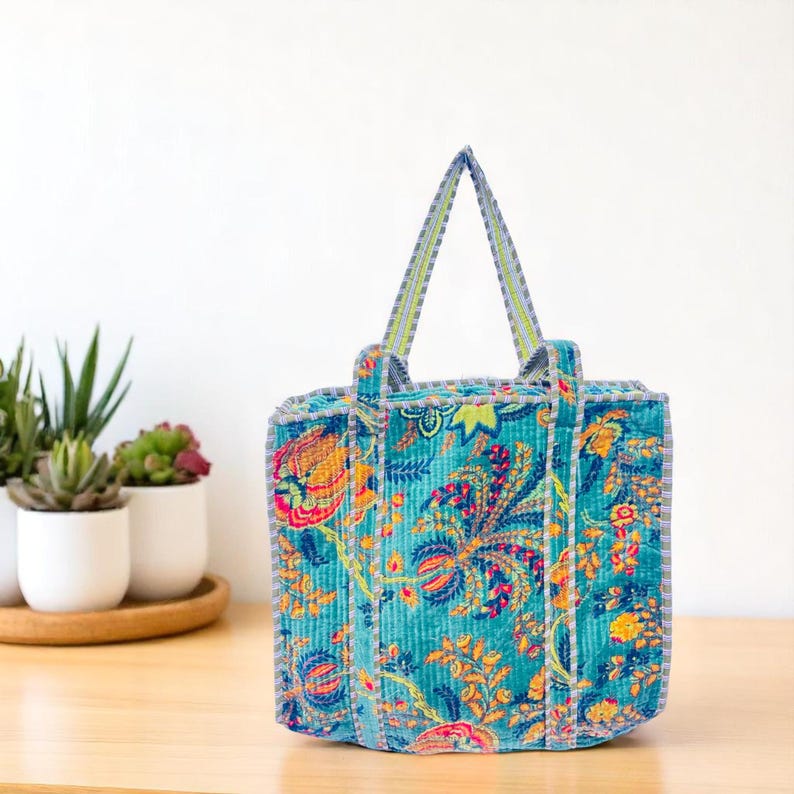 Colorful blue floral velvet shoulder tote bag placed on a wooden surface with potted plants in background.