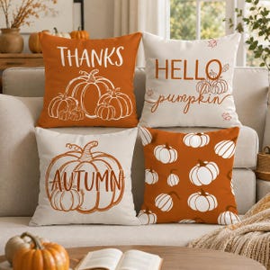 Pode incluir: Quatro almofadas quadradas com desenhos de abóboras. Uma é laranja com texto branco THANKS e contornos de abóboras brancas. Uma é branca com texto laranja HELLO pumpkin e folhas pequenas. Uma é branca com uma abóbora laranja e texto AUTUMN. Uma é laranja com um padrão de abóboras brancas.