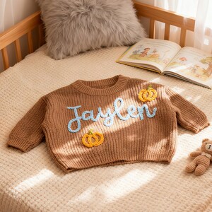 Op de afbeelding: Bruin gebreide trui met de naam "Jaylen" in lichtblauw schrift en twee gele pompoenapplicaties. De trui wordt gepresenteerd op een wieg met een pluche kussen en een boek. Een gehaakte pompoen en teddybeer zijn ook aanwezig.