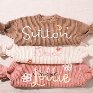 Pode incluir: Três camisolas de malha empilhadas, em tons de castanho, creme e rosa. Cada camisola apresenta nomes bordados: "Sutton", "Dudi" e "Cheryet Lollie". A camisola castanha tem detalhes de lua e estrela, as outras flores.
