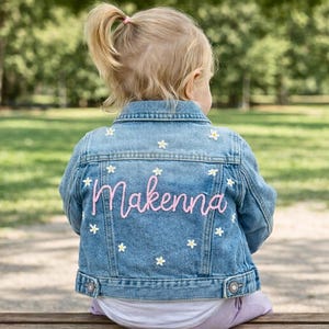 May include: A light blue denim jacket with the name "Makenna" embroidered in pink script on the back. The jacket is decorated with small white daisy flower appliqués. The child is sitting on a wooden bench.
