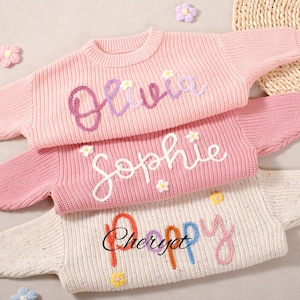 以下が含まれることがあります： パステルカラーのニットセーター3枚。紫色の「Olivia」、白の「Sophie」、様々な色の「Poppy」の名前が刺繍されています。各セーターはクルーネックと長袖で、小さな花の装飾が施されています。セーターは明るい背景に配置されています。