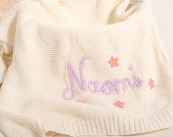 May include: Cream knitted blanket featuring the name "Naomi" embroidered in lavender script, with small pink and yellow flower details. The blanket has a ribbed edge.