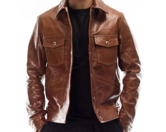 May include: A brown leather jacket with two chest pockets and a collared neckline. The jacket has a button-up front and is worn open, revealing a black shirt underneath. The leather has a textured appearance.
