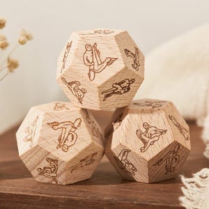 Könnte beinhalten: Drei Würfel aus Holz mit gravierten Figuren. Die Würfel sind auf einer Holzoberfläche gestapelt, mit einem Glas mit getrockneten Blumen und einem weißen Tuch im Hintergrund. Die Würfel haben eine natürliche Holzmaserung.
