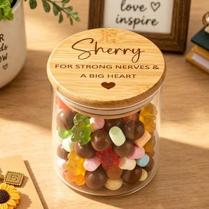 May include: A clear glass jar filled with assorted colorful gummy candies and chocolate balls, topped with a round bamboo lid engraved with the name Sherry and the text FOR STRONG NERVES & A BIG HEART with a small heart icon below the text.