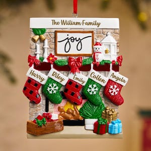 May include: A rectangular Christmas ornament featuring a fireplace design with five stockings, each bearing a name. The ornament includes the text "The William Family" and the word "joy". Red and green accents add festive color.