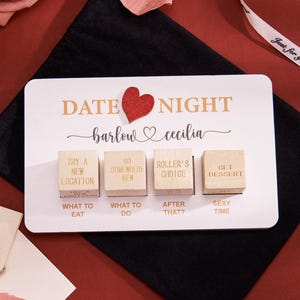 Może przedstawiać: Biała karta Date Night ze złotymi literami i czerwonym sercem. Cztery drewniane klocki z propozycjami aktywności znajdują się pod imionami "barlow" i "cecilia". Karta leży na czarnej powierzchni.