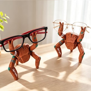 May include: Two brown, textured crab-shaped eyeglass holders made of a carved material. Each crab figure has articulated limbs with white stitched-like markings and teal accents, holding a pair of glasses aloft with its raised front claws.