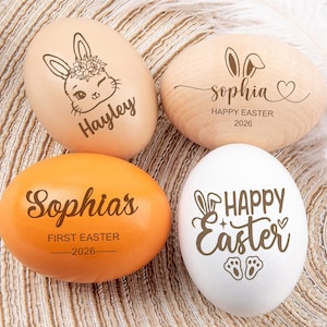 Könnte beinhalten: Vier dekorative Ostereier. Zwei sind hellbraun, eines ist orange und eines ist weiß. Jedes Ei hat ein einzigartiges Design, darunter Hasenohren, florale Akzente und die Worte "Hayley", "Sophia", "Happy Easter 2026" und "Sophia's First Easter 2026".