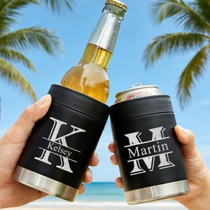 Könnte beinhalten: Zwei schwarze isolierte Getränkehalter mit silbernen Böden werden zum Anstoßen hochgehalten. Einer hält eine Bierflasche, der andere eine Dose. Jeder hat ein weißes Monogramm und den Namen: "Kelsey" und "Martin". Palmen und blauer Himmel im Hintergrund.
