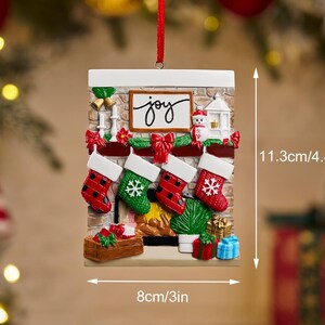 May include: Christmas ornament shaped like a fireplace with four stockings, measuring 4.4 inches tall and 3 inches wide. The ornament has the word "joy" and a snowman. The stockings are red and green with white cuffs.