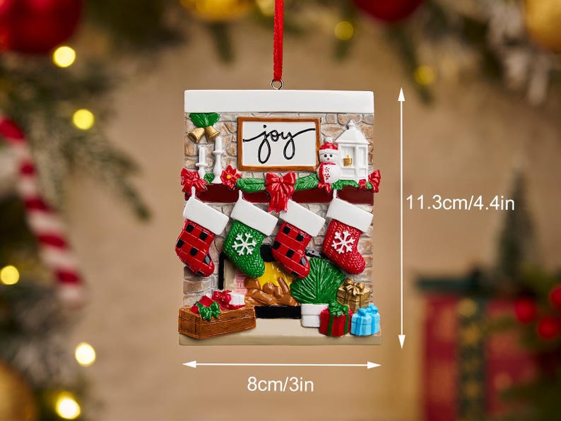May include: Christmas ornament shaped like a fireplace with four stockings, measuring 4.4 inches tall and 3 inches wide. The ornament has the word "joy" and a snowman. The stockings are red and green with white cuffs.