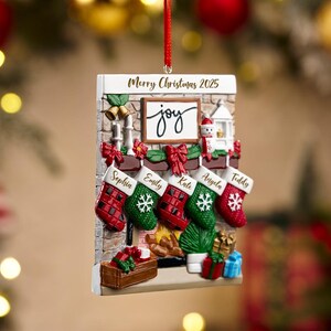 May include: A Christmas ornament shaped like a fireplace with stockings. The ornament has the words "Merry Christmas 2025" and "joy". Stockings are personalized with names. Red ribbon for hanging.