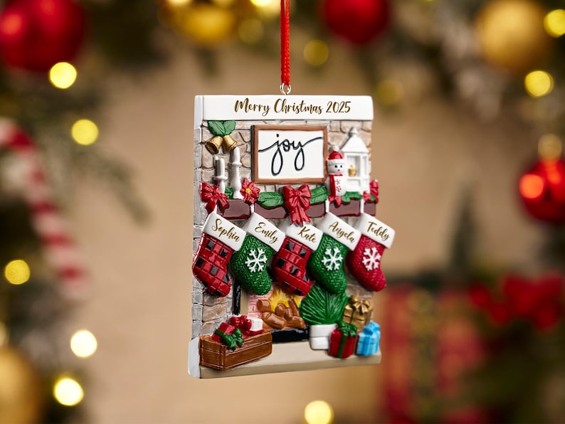 May include: A Christmas ornament shaped like a fireplace with stockings. The ornament has the words "Merry Christmas 2025" and "joy". Stockings are personalized with names. Red ribbon for hanging.