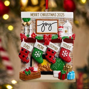 May include: Christmas ornament shaped like a fireplace with four stockings labeled Dad, Mom, Sophia, and Teddy. The ornament has the text "Merrt Chrustmas 2025" and a sign that says "joy". Red and green accents and gifts complete the festive scene.