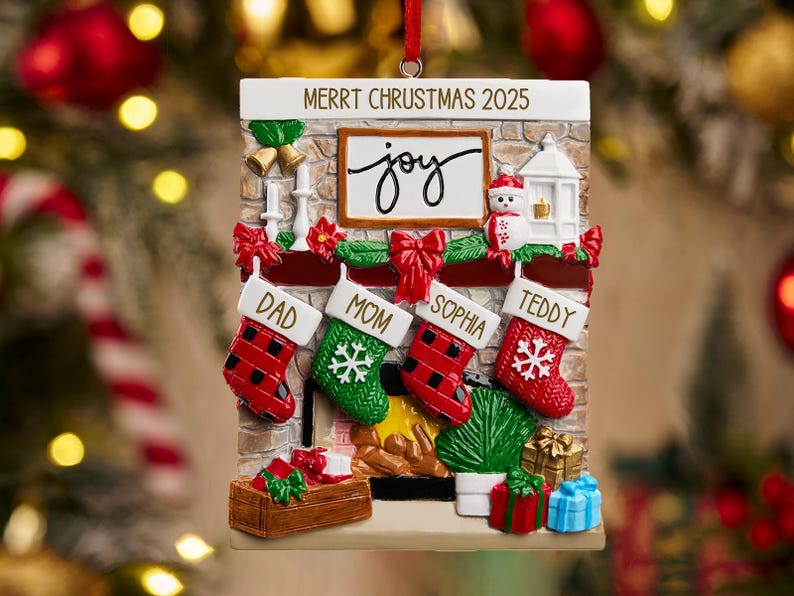 May include: Christmas ornament shaped like a fireplace with four stockings labeled Dad, Mom, Sophia, and Teddy. The ornament has the text "Merrt Chrustmas 2025" and a sign that says "joy". Red and green accents and gifts complete the festive scene.