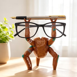 May include: A brown, crab-shaped desk organizer with teal accents and white stitch-like markings. The figure holds a pair of black-framed eyeglasses in its raised claws, while a metallic gold and black fountain pen rests across the top.