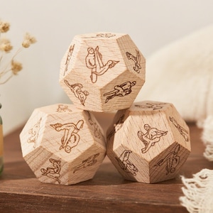 May include: Three wooden dodecahedron dice with engraved figures. The dice are stacked on a wooden surface, with a glass jar of dried flowers and a white cloth in the background. The dice are made of light-colored wood.