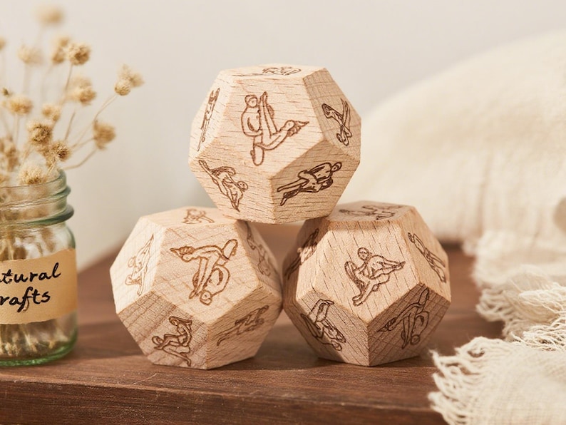 May include: Three wooden dodecahedron dice with engraved figures. The dice are stacked on a wooden surface, with a glass jar of dried flowers and a white cloth in the background. The dice are made of light-colored wood.