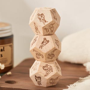 May include: Three wooden, dodecahedron-shaped blocks stacked on top of each other. Each block is engraved with various illustrations of human figures. The blocks are light brown and rest on a dark brown wooden surface.