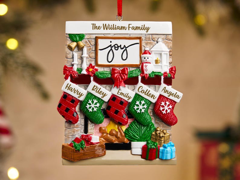 May include: A rectangular Christmas ornament featuring a fireplace design with five stockings, each bearing a name. The ornament includes the text "The William Family" and the word "joy". Red and green accents add festive color.